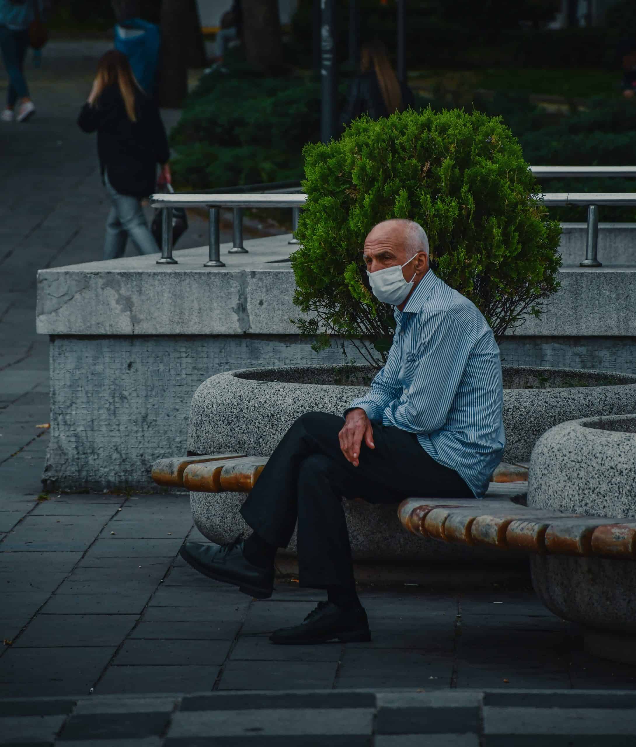 En eldre mann sitter på en benk og har på seg et munnbind