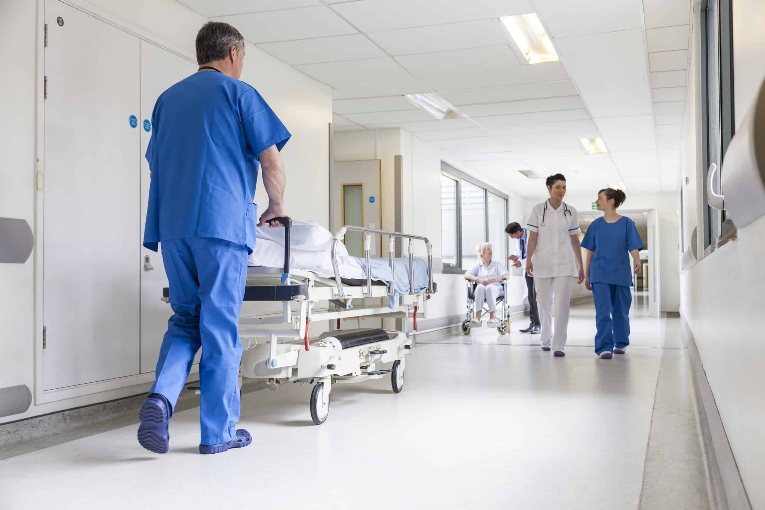 Sykehuskorridor med mannlig sykepleier som triller en sengepasient til behandling, andre sykepleiere i bakgrunnen