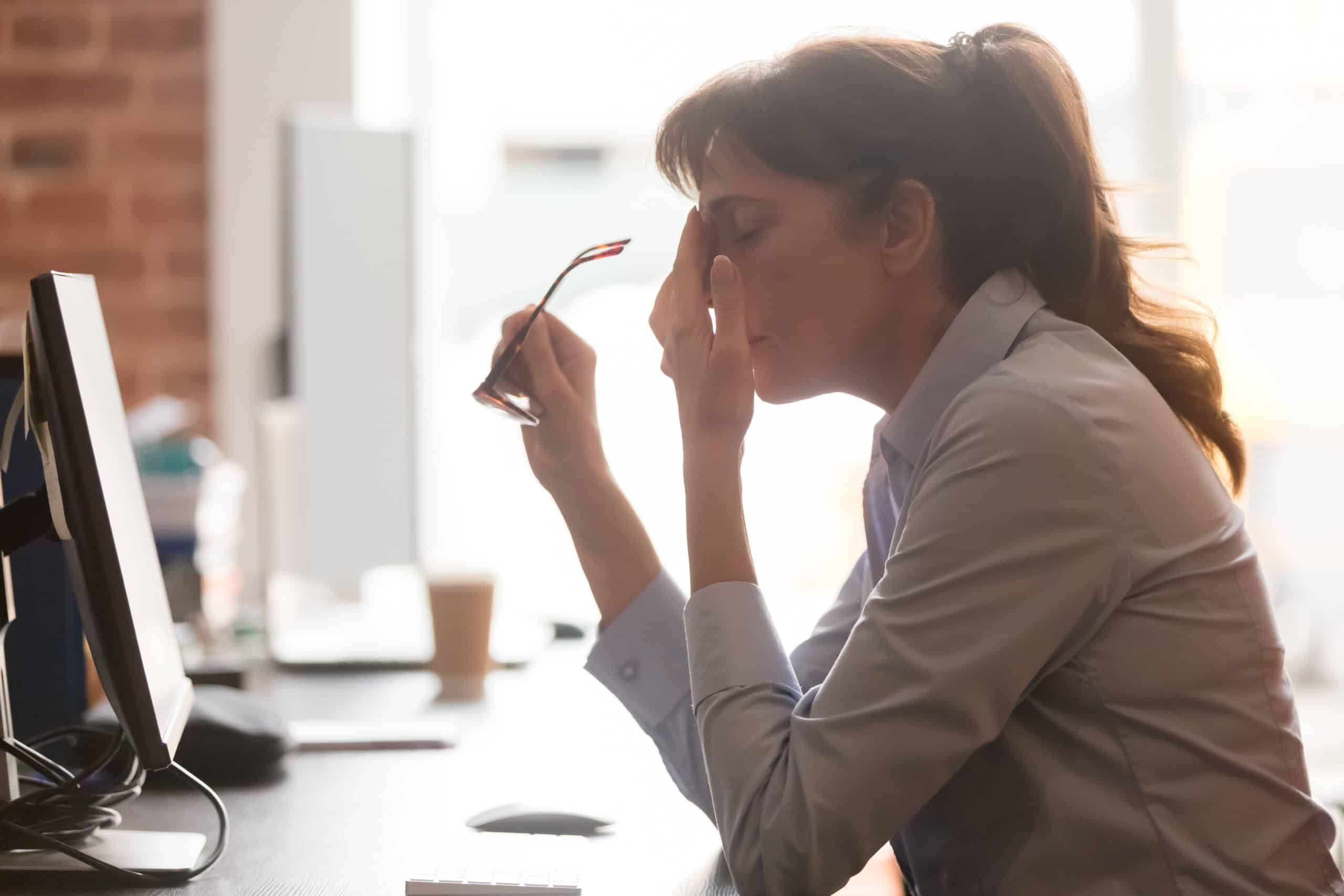 Utmattet kvinnelig arbeidstaker sitter ved kontorpulten og tar av briller